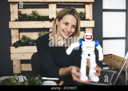 Hübsche junge blonde Frau sitzt in einem nachhaltigen Büro und Funktioniert mit einem kleinen Roboter Stockfoto