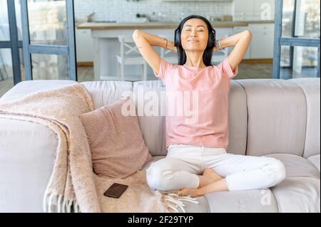Junge Erwachsene asiatische Frau in kabellosen Kopfhörern und dehnbare Kleidung, sitzen in Yoga-Position auf der Couch mit den Händen hinter dem Kopf, meditieren zu ihrer Lieblings-ruhige Musik, hören sie aus Telefon Stockfoto