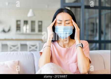 Junge asiatische Frau mit geschlossenen Augen in schützende Gesichtsmaske sitzen auf der Couch zu Hause, berühren Tempel, leiden an Migräne oder schweren Kopfschmerzen, krank fühlen, mit Symptomen der Coronavirus-Grippe Stockfoto
