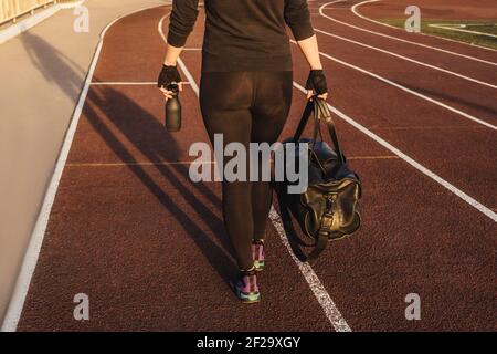 Fitness Frau in schwarz Trainingsanzug im Freien, Stadion Tracks Hintergrund. Frau mit Sporttasche geht vom Training weg. Abendlicht. Stockfoto