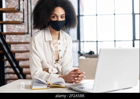 Multirassische Geschäftsfrau in schwarzer Maske im Büro am Laptop, Gefahr der Infektion des Virus Coronavirus-Infektion, Pandemie in der Welt, Schutz Mitarbeiter am Arbeitsplatz, Sicherheit des Personals. Stockfoto