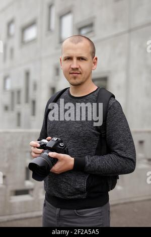 Lächelnd schöner männlicher Fotograf mit moderner dslr-Kamera posiert über Betongebäude im Hintergrund Stockfoto