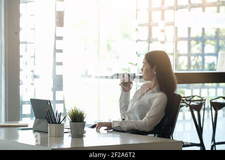 Portrait einer schönen Frau oder Buchhalterin, die am Schreibtisch sitzt Modernes Büro mit Innen trinken Heißgetränk halten Tasse mit Kaffee in den Händen Stockfoto