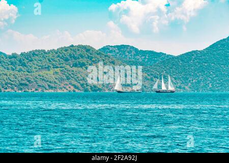 Sommerkonzept: Zwei weiße Segelyachten, die auf dem blauen Wasser der Ägäis vor einer grünen und felsigen Waldinsel schweben.natürlicher Hintergrund Stockfoto