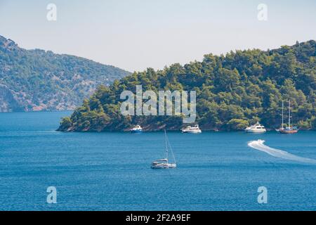 Sommerkonzept: Eine weiße Segelyacht schwimmt auf dem blauen Wasser der Ägäis. Viele Yachten vor einer grünen Waldinsel vor Anker. Stockfoto