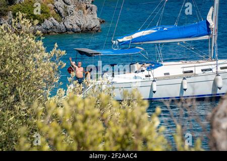 Hinter einem grünen Baum ein einzelnes Segelschiff mit einem Mann. Die Person trägt Badeshorts und winkt der Kamera zu. Blaues Wasser der Ägäis Stockfoto