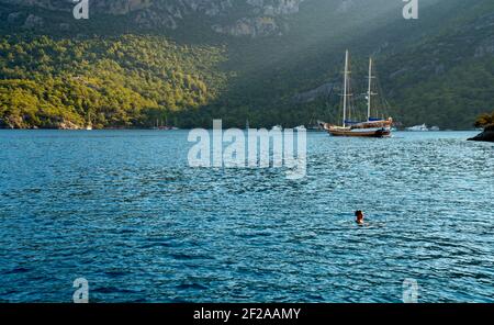 Sommerkonzept: Eine türkische Gulet Yacht, die an der Ägäis mit Sonnenstrahl im Hintergrund verankert ist. Eine alleinstehende Frau, die im See schwimmend ist. Grün - blauer Kontrast Stockfoto