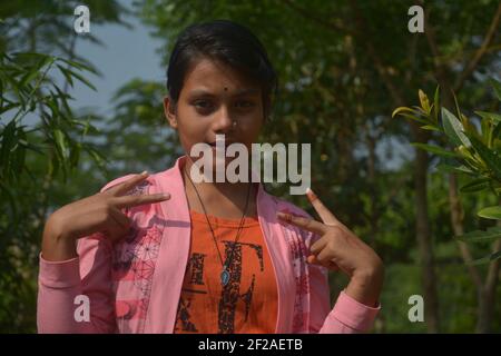 Close up schöne indische Bengali Teenager-Mädchen trägt rosa Kleid, Schnur Medaillon, Nosering, schwarze Bindi auf der Stirn, Haarbeuge, zeigt Siegeszeichen Stockfoto