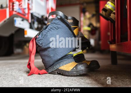 Feuerwehrschuhe und -Hosen und ein Schrank, Feuerwehrauto und Feuerwehrmann im Hintergrund. Stockfoto