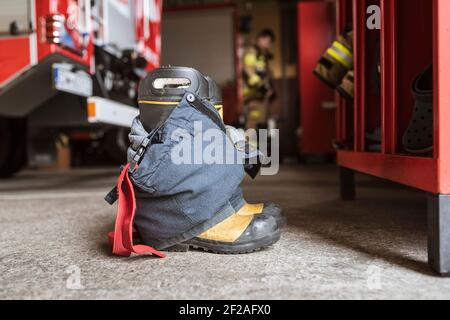 Feuerwehrschuhe und -Hosen und ein Schrank, Feuerwehrauto und Feuerwehrmann im Hintergrund. Stockfoto