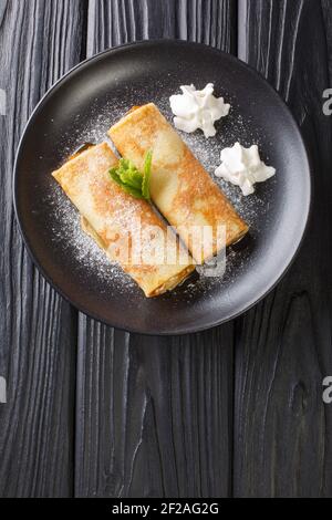 Leckere Crepes mit Aprikosenmarmelade, Schlagsahne und Puderzucker aus nächster Nähe auf einem Teller auf dem Tisch. Vertikale Draufsicht von oben Stockfoto