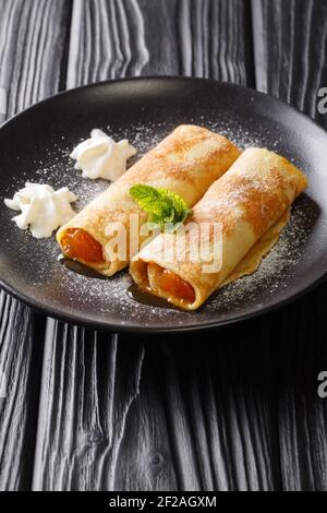 Leckere Crepes mit Aprikosenmarmelade, Schlagsahne und Puderzucker in Nahaufnahme auf einem Teller auf dem Tisch. Vertikal Stockfoto