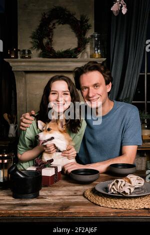 Glückliches kaukasisches Paar, das in der Küche mit ihrem Corgi-Hund kocht. Junge Familie, Frau und Mann essen zu Abend Stockfoto