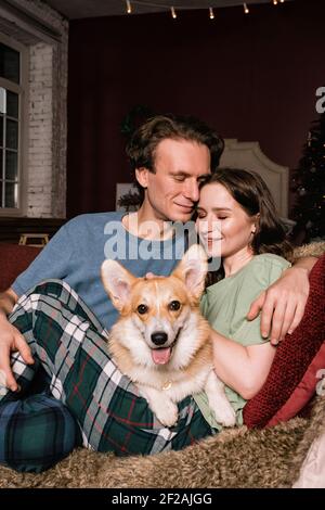 Glückliches kaukasisches Paar auf dem Sofa mit ihrem Corgi Hund. Junge Familie, Frau und Mann essen zu Abend Stockfoto