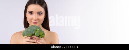 Banner – Langformat. Porträt einer jungen schönen Brünette, kaukasische Frau hält grünen Brokkoli in den Händen, isoliert auf grauem Hintergrund mit Stockfoto