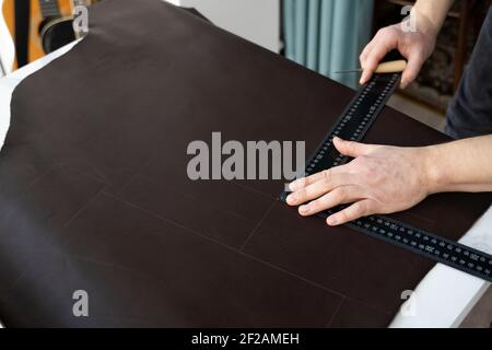 Herrenhand, die eine Ahle und ein Metalllineal hält und an einer Ledertasche in seiner Werkstatt arbeitet. Arbeitsprozess mit einem braunen Naturleder. Handwerker Stockfoto