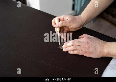 Herrenhand, die eine Ahle und ein Metalllineal hält und an einer Ledertasche in seiner Werkstatt arbeitet. Arbeitsprozess mit einem braunen Naturleder. Handwerker Stockfoto