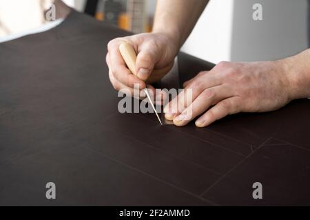 Herrenhand, die eine Ahle und ein Metalllineal hält und an einer Ledertasche in seiner Werkstatt arbeitet. Arbeitsprozess mit einem braunen Naturleder. Handwerker Stockfoto