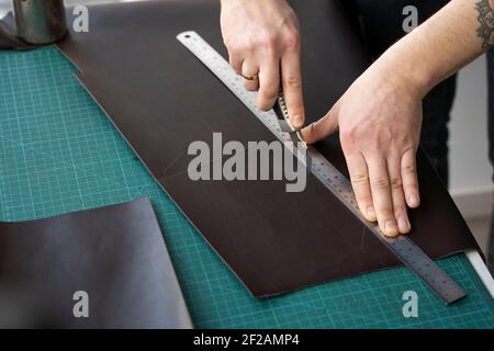 Herrenhand hält ein Schreibwarenmesser und ein Metalllineal und schneidet auf einem Stück für eine Ledertasche in seiner Werkstatt. Arbeitsprozess mit einem braunen Stockfoto