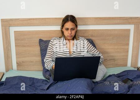 Junge Frau, Mitte dreißig, Telearbeit mit ihrem Laptop, sitzend und mit einer blauen Bettdecke bedeckt, in der Bequemlichkeit ihres Schlafbettes. Stockfoto