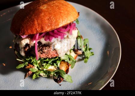 Frischer saftiger Cheeseburger mit Rucola und roten eingelegten Zwiebeln auf einem hausgemachten Brötchen auf einem hellblauen Vintage-Teller. Nahaufnahme, Draufsicht. Stockfoto