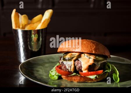 Klassischer saftiger Burger mit Tomatenscheiben, Käse und eingelegten Gurken. Der Metalleimer mit pommes Frites in der Nähe des Tellers. Stockfoto