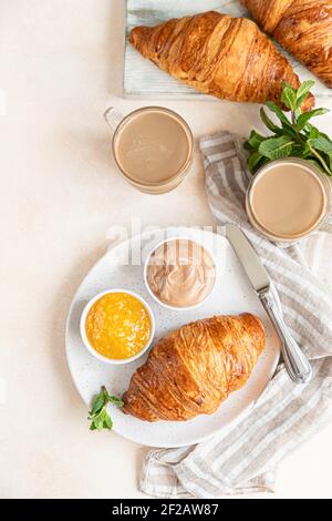 Frisch gebackene knusprige französische Croissants mit Marmelade und Schokoladencreme und zwei Kaffeetassen, heller Betonhintergrund. Frühstück. Selektiver Fokus. Oben vi Stockfoto