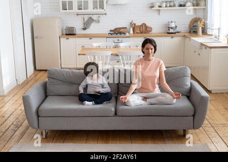 Mutter macht Yoga-Übung, sitzen auf der Couch zu Hause, Kind mit Gadget, spielen ein Spiel auf Tablet. Stockfoto