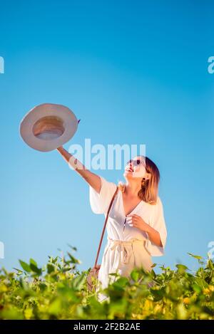 Fröhlich stilvolle junge Frau mit Hut in der Hand in Offenes Sommerfeld lächelnd Stockfoto