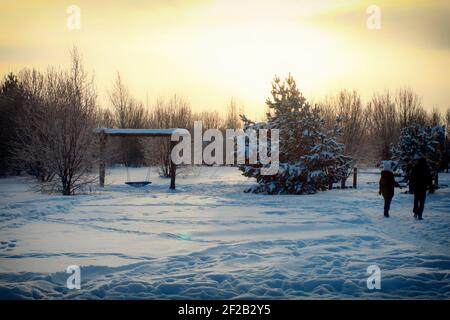 Winterlandschaft bei Sonnenuntergang mit großen Schaukeln. Die Menschen gehen den Weg entlang in den Wald. Goldene Abendsonne untergeht hinter den verschneiten Bäumen. Stockfoto