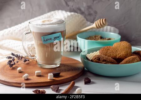 Kaffee Latte Tasse mit Love You Aufkleber auf Holztablett Mit türkisfarbenen Schalen mit Plätzchen und Marshmallows an der grauen Wand Hintergrund Stockfoto