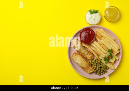 Teller mit gebratenem Fisch und Pommes, grünen Erbsen und Saucen auf einem leuchtend gelben Hintergrund, lebhafter Pub-Essen Stockfoto