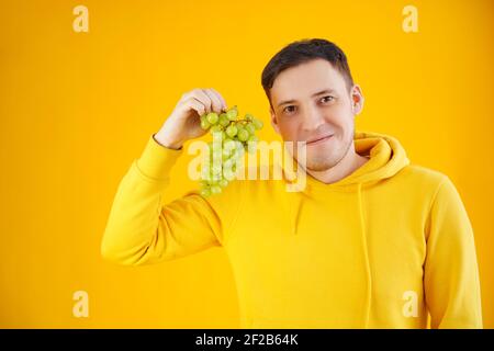 Porträt eines jungen Mannes mit grünen Trauben auf gelbem Hintergrund. Nahaufnahme von schönen Kerl in gelben Kapuzenpullover hält Trauben Stockfoto