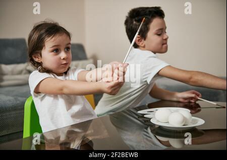 Zwei charmante Kinder halten Pinsel bereit, um Ostereier mit Farben zu dekorieren. Stockfoto