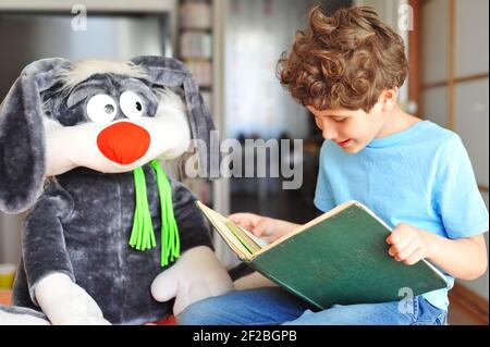 Bezaubernder kleiner Junge mit lockigem Haar liest ein aufregendes, lustiges und Interessantes Buch zu seinem Plüschkaninchen und lächelt Stockfoto