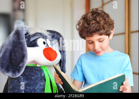 Bezaubernder kleiner Junge mit lockigem Haar liest ein aufregendes, lustiges und Interessantes Buch zu seinem Plüschkaninchen und lächelt Stockfoto