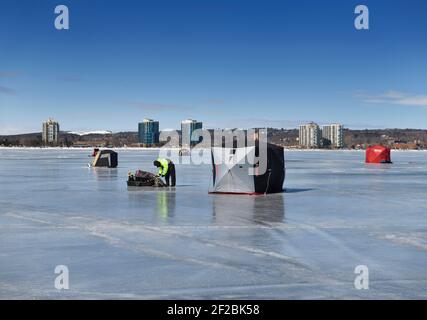 Barrie, Ontario, Kanada - 7. März 2021: Fischer packen ein Eis Angeln Zelt auf gefrorenen Kempenfelt Bay of Lake Simcoe im Winter mit Barrie Eigentumswohnungen Stockfoto