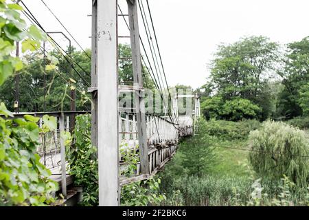 Alte eiserne Hängebrücke, die nicht funktioniert, über eine getrocknete Flussseite. Auf dem gegenüberliegenden Ufer Weide und Fichte. Vintage. Stockfoto