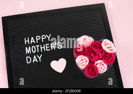 Schwarzer Briefbogen mit weißen Plastikbuchstaben mit Zitat Happy Mothers Day und Blumenseife, Herz auf rosa Hintergrund. Stockfoto
