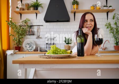 Entspannte junge Frau posiert, am Küchentisch stehend. Erwachsene Brünette Ruhe mit Alkohol und Obst in der Küche Stockfoto