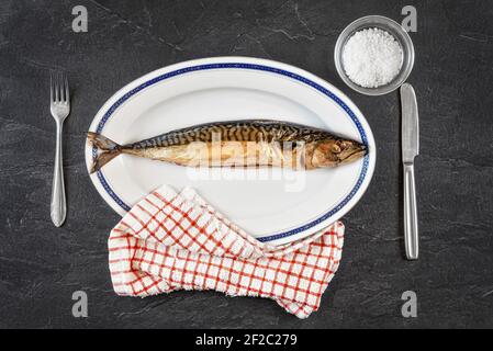 Ganze geräucherte Makrelenfische auf weißem ovalem Teller, grauschwarzer Stein wie Tischunterwand. Blick von oben Stockfoto