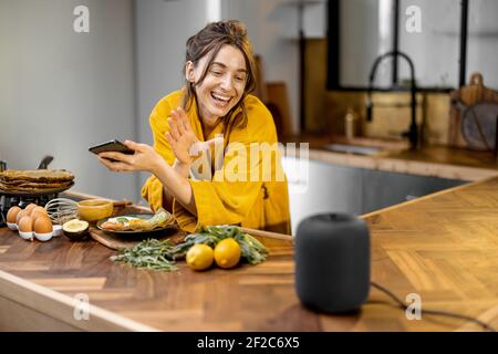 Glückliche Frau im Bademantel Steuerung Heimgeräte mit einer Sprachbefehle, sprechen zu einer intelligenten Spalte während der Frühstückszeit in der Küche zu Hause. Smart Home-Konzept Stockfoto