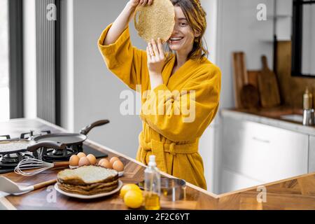 Porträt einer jungen fröhlichen Frau, die ihr Gesicht bedeckt Gebratener Pfannkuchen beim Kochen Frühstück in der Küche zu Hause Stockfoto