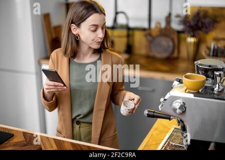 Junge stilvolle Frau legte die Tasse neben der Kaffeemaschine. Kaffee trinken, bevor Sie morgens in der schönen Küche arbeiten und sich am Telefon unterhalten. Stockfoto