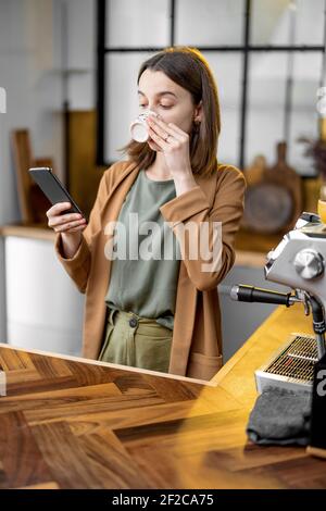 Pingelige junge Frau in Eile, um am Morgen zu arbeiten, tragen Jacke, trinken Kaffee und plaudern am Telefon unterwegs. Tägliche Routine der Geschäftsfrau. Auf das Telefon schauen. Stockfoto