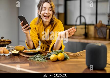 Glückliche Frau im Bademantel Steuerung Heimgeräte mit einer Sprachbefehle, sprechen zu einer intelligenten Spalte während der Frühstückszeit in der Küche zu Hause. Smart Home-Konzept Stockfoto
