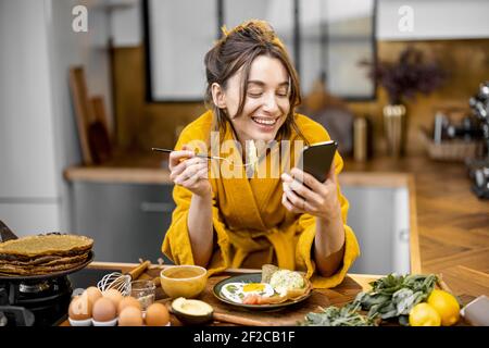 Junge glückliche Frau in gelben Bademantel gekleidet genießt gesundes Frühstück und Lesung am Telefon zu Hause. Morgen Angelegenheiten und Routine Stockfoto