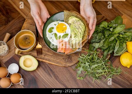 Gesundes Frühstück aus Ei, in Scheiben geschnittener Avocado, gesalzenem rotem Fisch und Pfannkuchen auf grünem Teller auf einem hölzernen Küchentisch, Blick von oben Stockfoto