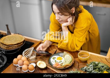 Junge glückliche Frau in gelben Bademantel gekleidet genießt gesundes Frühstück und Lesung am Telefon zu Hause. Morgen Angelegenheiten und Routine Stockfoto