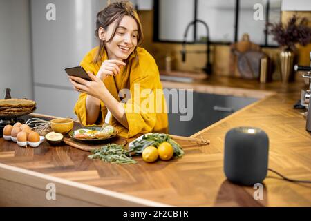 Glückliche Frau im Bademantel Steuerung Heimgeräte mit einer Sprachbefehle, sprechen zu einer intelligenten Spalte während der Frühstückszeit in der Küche zu Hause. Smart Home-Konzept Stockfoto